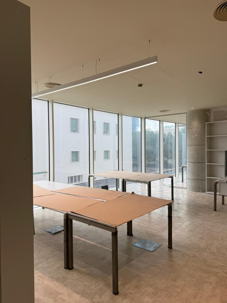 Modern office with large windows and desks covered in brown paper under linear lighting.