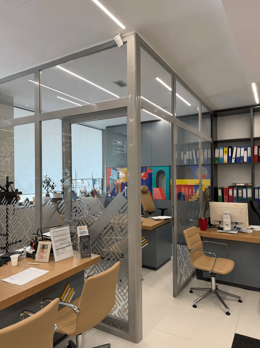 Modern office featuring glass partitions, wooden desks, tan chairs, and shelves with colorful binders.
