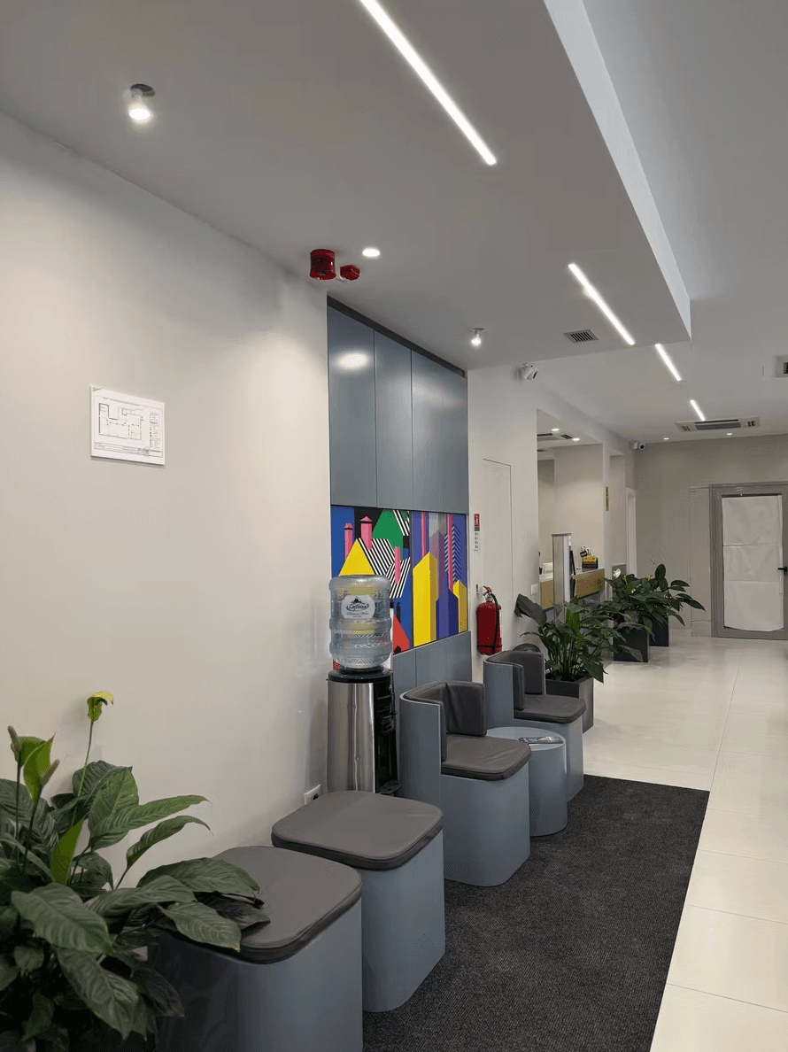 Modern office waiting area featuring gray stools, a water cooler, colorful art, and potted plants.